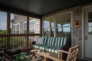 Une véranda couverte avec un canapé en osier, des meubles en bois et de grandes fenêtres donnant sur une maison.