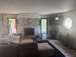 A living room with a fireplace, a sofa, a TV, and a window with a view of trees. - 3-bedroom house in soothing Anchorage perfect for your getaway (Anchorage)