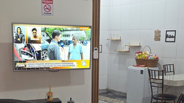 A room with a television displaying a news program, black wicker chairs, a red rug, a small table with a basket, and a white tiled wall. - Studio apartment near the shopping mall. (Campo Grande)