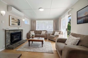 A living room with a fireplace, sofas, a coffee table, and a painting on the wall. - Red Door 56 - Ride to Redwoods, Sleeps 4 (Rotorua)