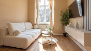 Un salón moderno con sofá, una pequeña mesa redonda con una planta, un televisor montado en la pared y una ventana con cortinas.