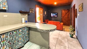 A room with a kitchen area, a seating area with a sofa, a television mounted on the wall, and a wooden door. - 🌋Near La Fortuna : Private villa with private pool surrounded by nature (Provincia de Alajuela)