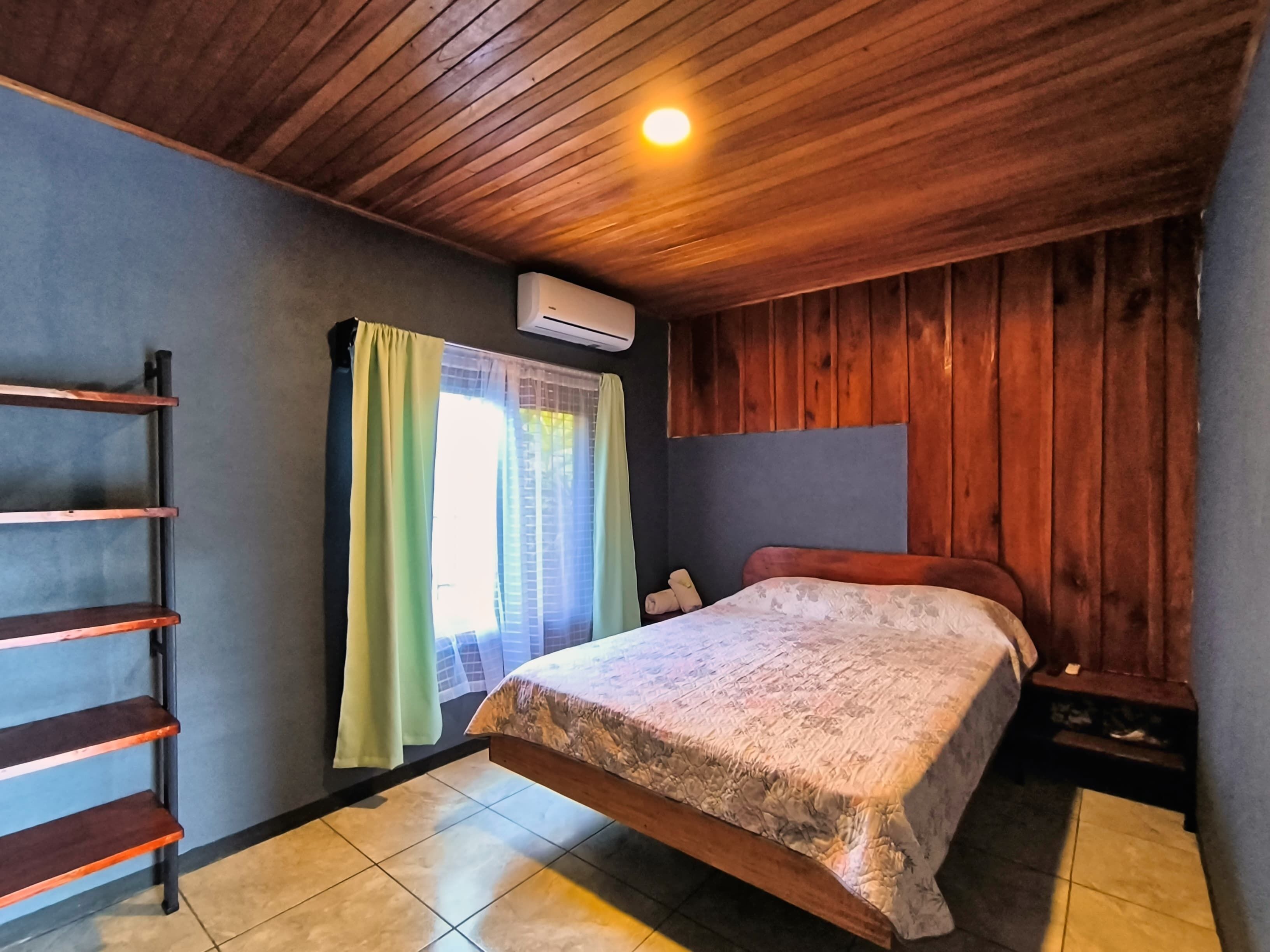 Un dormitorio con cama, cabecera de madera, ventana con cortinas, aire acondicionado de pared y estante de madera.