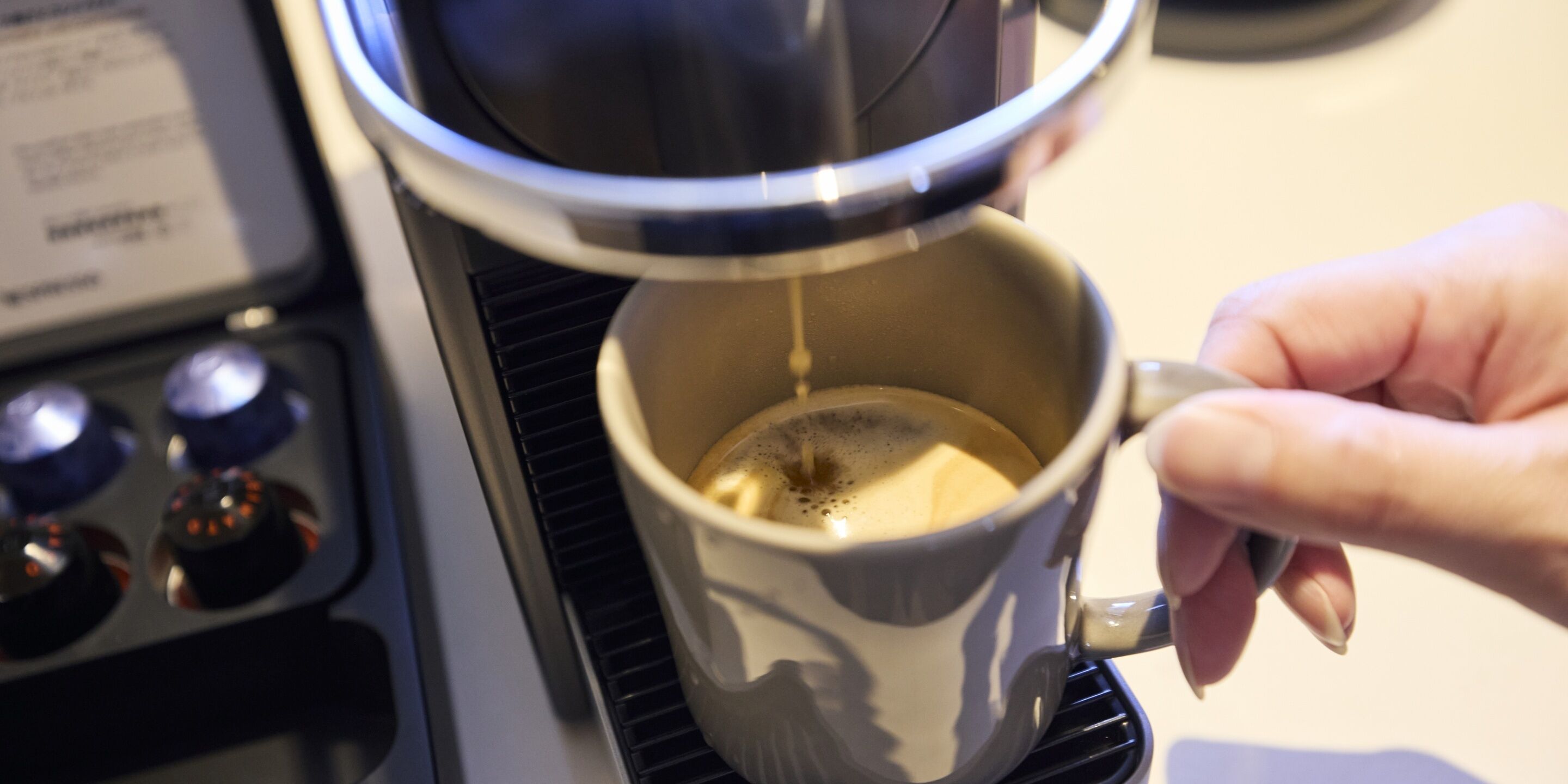A hand holding a mug under a coffee machine, with steam rising from the cup.