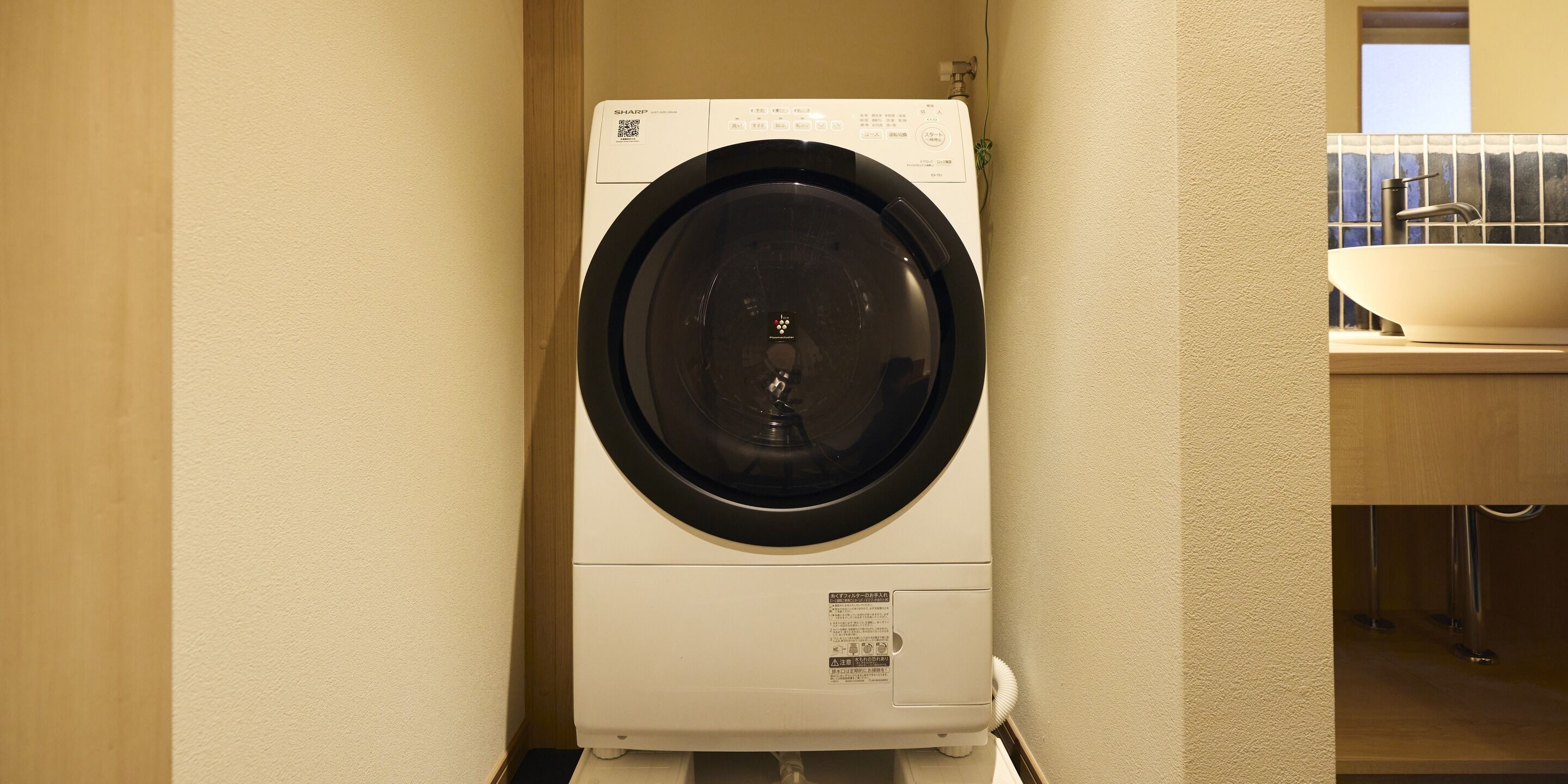 A compact, white front-loading washing machine in a small laundry room with a sink and a window.