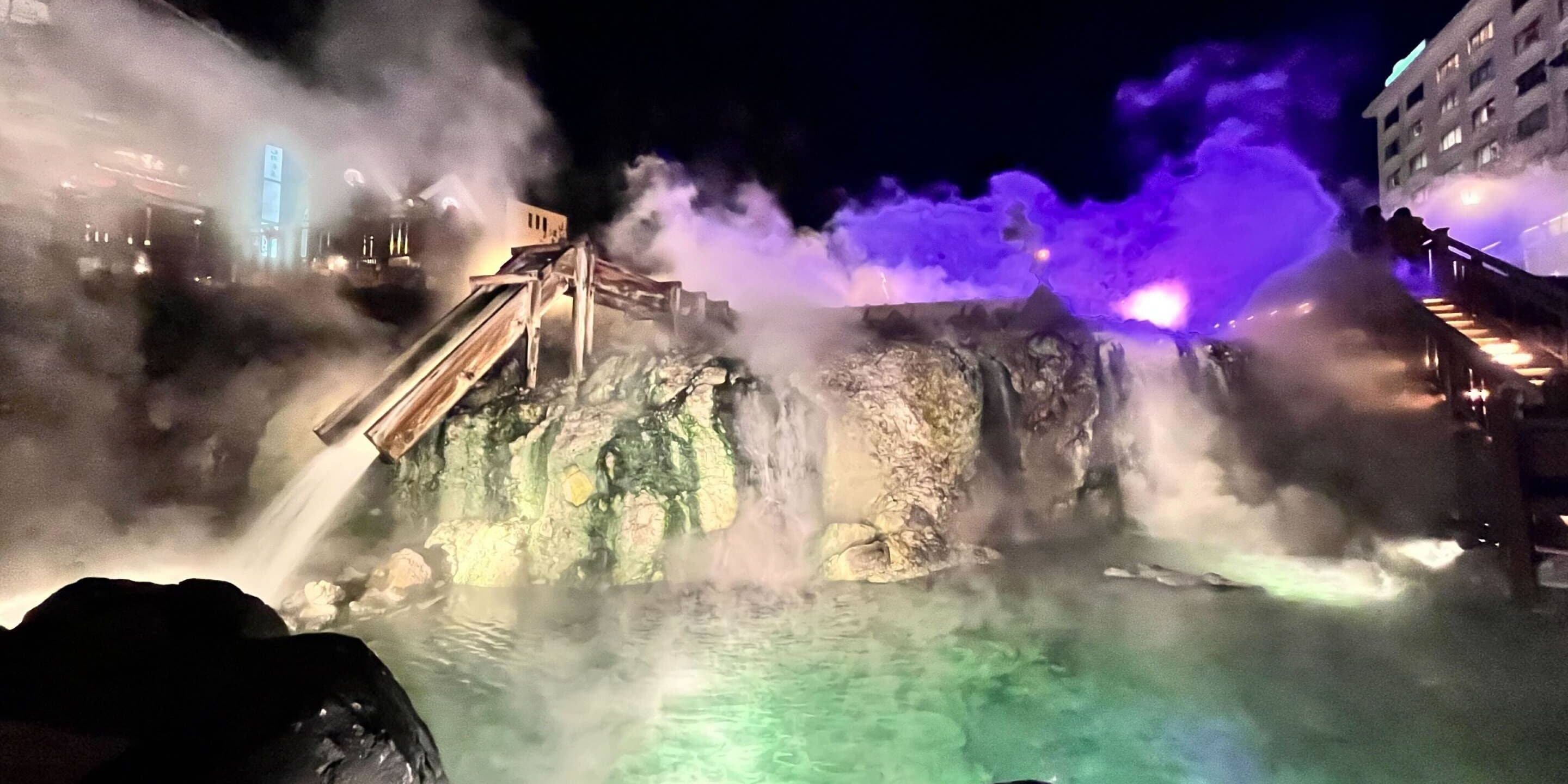 A hot spring area with a wooden walkway, steam, and a building in the background.