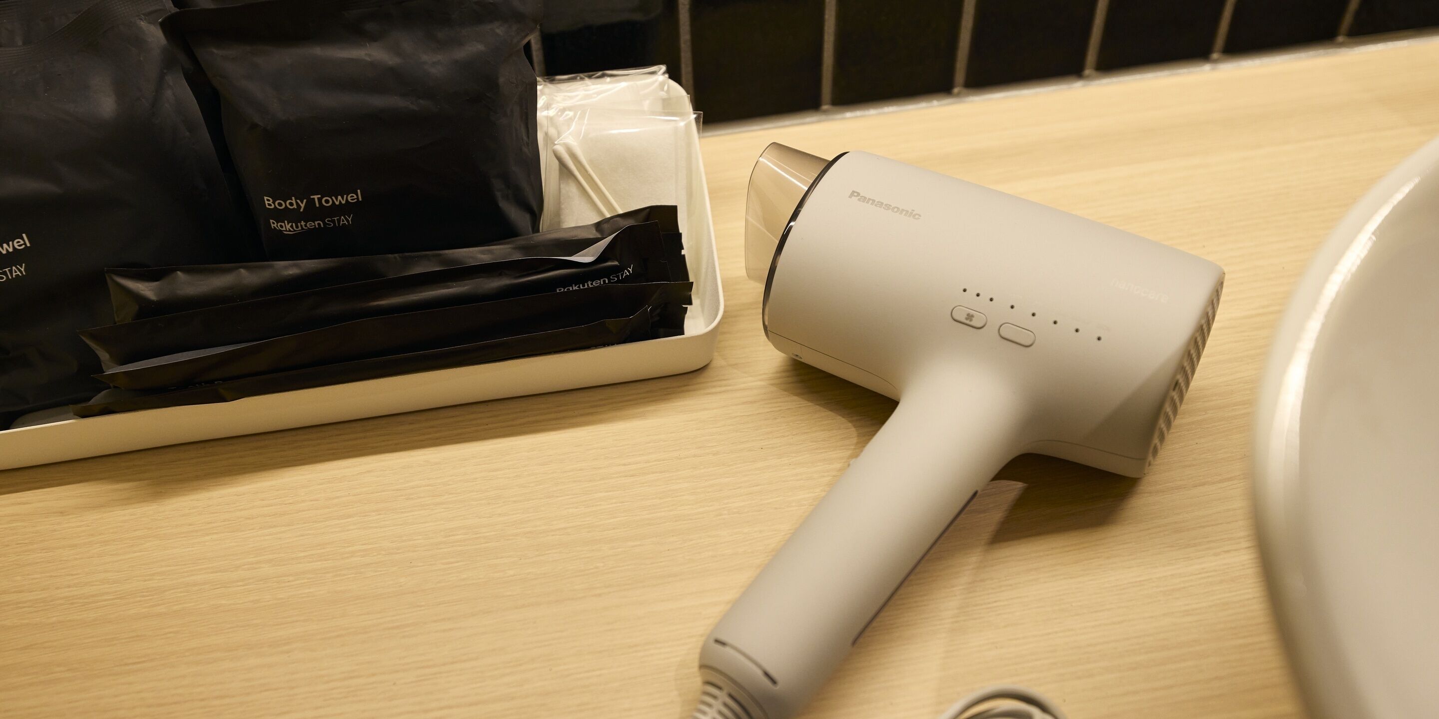 A Panasonic hair dryer on a wooden surface with a plastic container and black bags labeled 'Body Towel Rakuten STAY'.