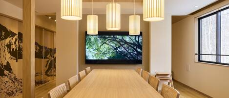 A conference room with a long table, four chairs, a large screen, and hanging pendant lights.