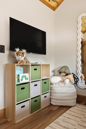A room with a wooden floor, a bookshelf with green and white storage bins, a plush toy on top, a television mounted on the wall, and a large mirror with a wavy frame.