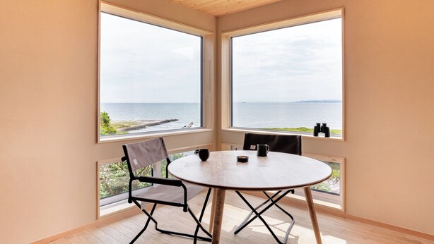 Une pièce avec une table ronde et deux chaises, de grandes fenêtres donnant sur la mer et un plafond en bois.