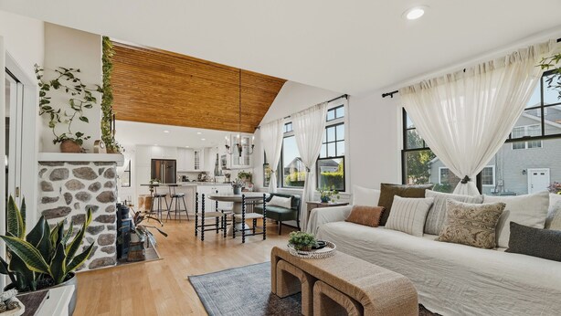 A modern living room with a stone fireplace, a wooden ceiling, a large window with curtains, a sofa, and a dining area with chairs.