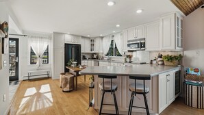 A modern kitchen with white cabinetry, a central island with stools, a built-in microwave, and a window with curtains.