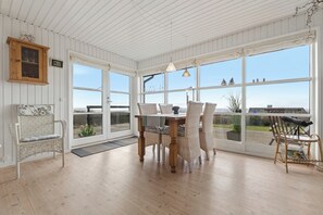 Una habitación con una mesa y sillas de madera, un banco de mimbre y ventanas grandes con vistas.