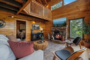 A log cabin interior with a fireplace, wooden furniture, a balcony, and a large window. - *New Panoramic Smoky Mtn Views-Mins to Dollywood (Sevierville)