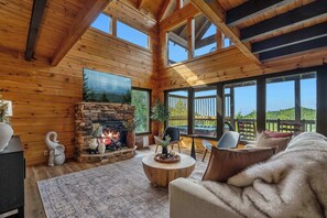 L’intérieur chaleureux d’un chalet, avec une cheminée en pierre, des murs en bois et de grandes fenêtres offrant une vue sur l’extérieur.