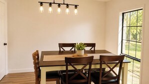 A dining area with a wooden table and chairs, a hanging light fixture, and a view through a window.