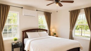 A bedroom with a bed, a ceiling fan, an air conditioning unit, a nightstand with a lamp, and a window with curtains.