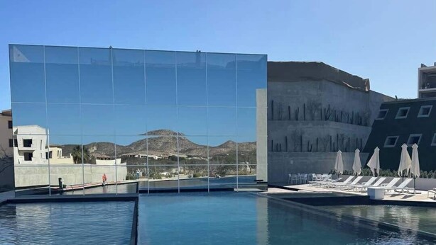 En pool med solstolar och parasoller, en glasvägg som speglar ett bergigt landskap och en byggnad med modern design.