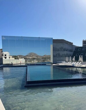 A swimming pool with lounge chairs and umbrellas, a glass wall reflecting a mountainous landscape, and a building with a modern design. - New Golf Course Condo with Ocean Views (San José del Cabo)