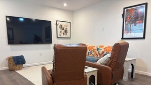 Un salon doté d’un téléviseur à écran plat, de deux fauteuils en cuir marron, d’une petite table blanche, d’un panier en osier et d’une œuvre d’art encadrée accrochée au mur.