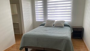 Makuuhuone, jossa on sänky, ikkuna, jossa on sälekaihtimet, puulattia ja pieni yöpöytä.