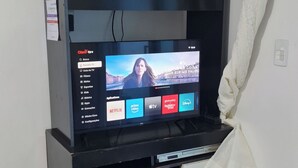 A TV stand with a flat-screen TV displaying a TV guide interface. - Entire house 350m from Ingleses Beach, sleeps up to 5 people. (Florianópolis)