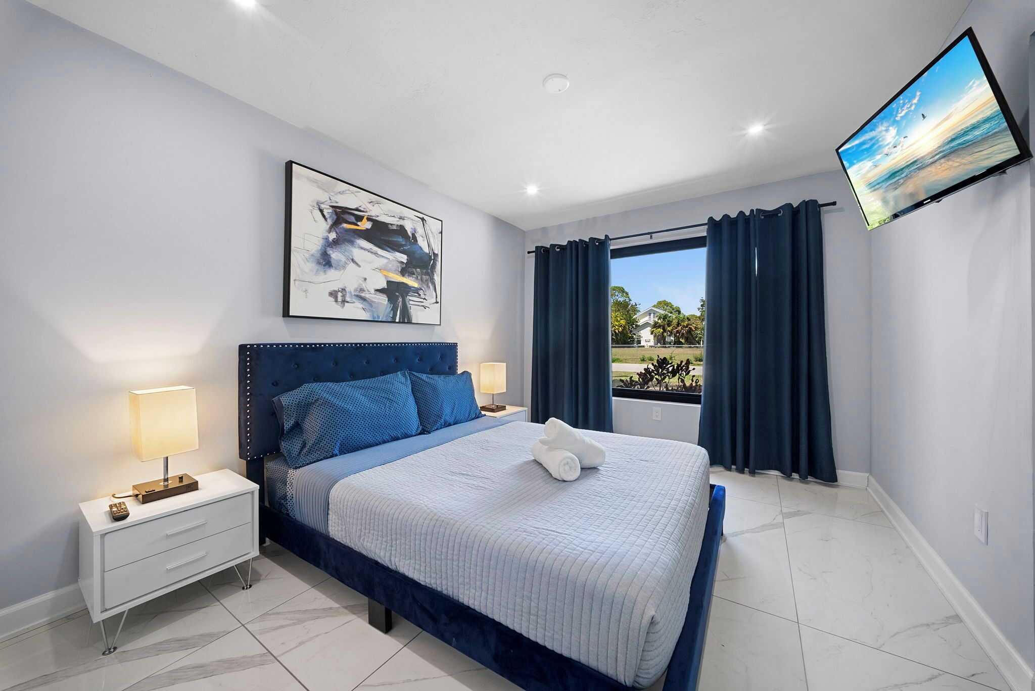 A bedroom with a bed, bedside tables, a TV, a window with curtains, and a painting on the wall.