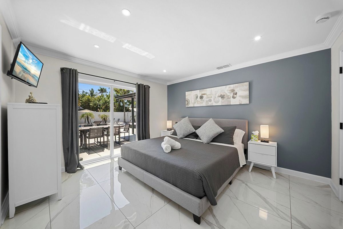 A modern bedroom with a large bed, a flat-screen TV mounted on the wall, and a sliding glass door leading to an outdoor area.