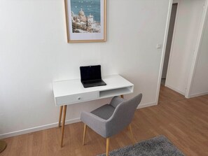 Un bureau blanc avec un ordinateur portable, une chaise grise et, au mur, un tableau représentant une ville côtière.