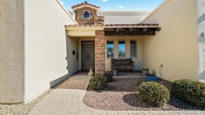 Un edificio residenziale con tetto in tegole, un piccolo camino in mattoni, un ingresso con cancello e un patio pavimentato con due cespugli e un tubo da giardino.
