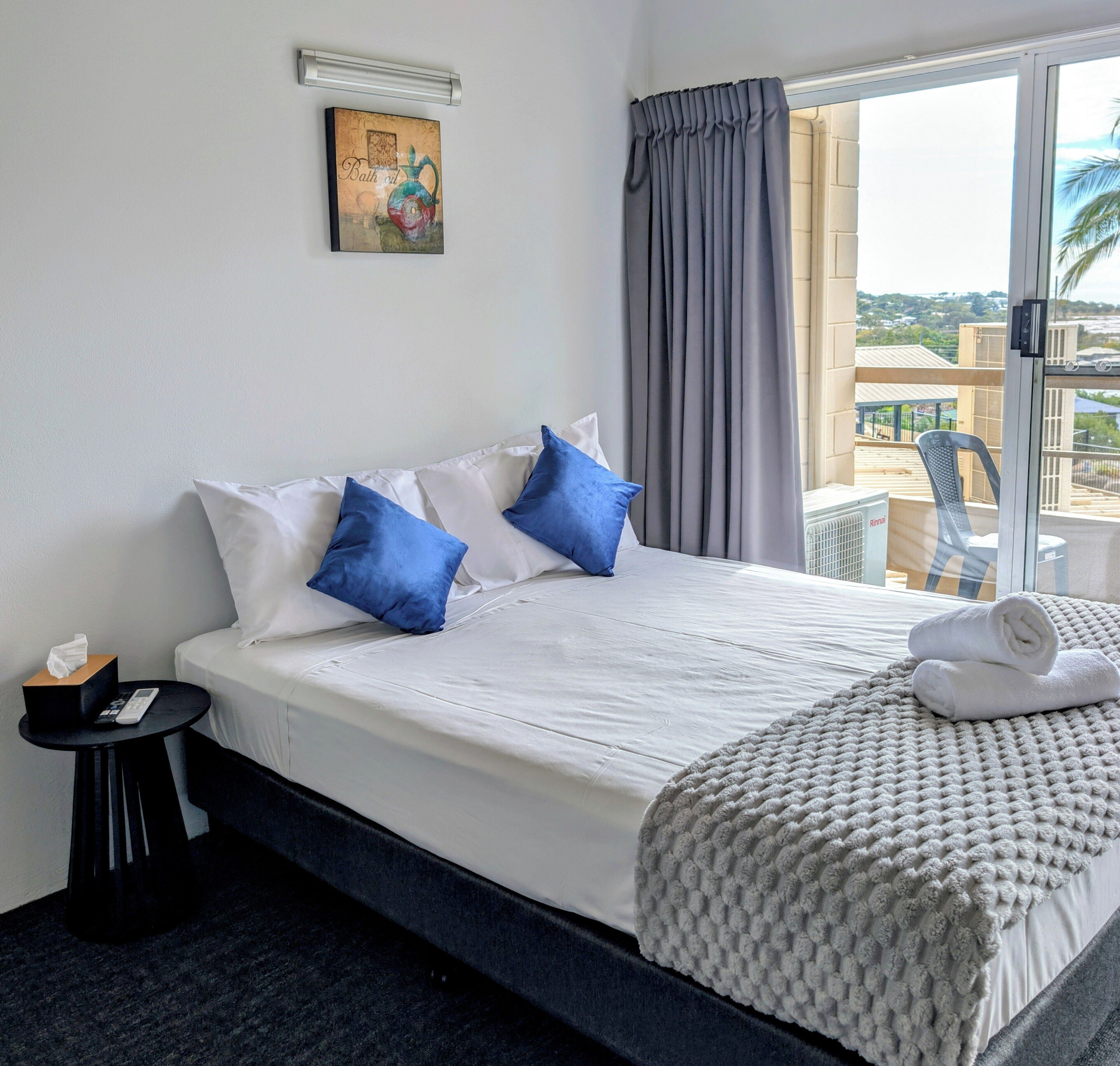 Une chambre d’hôtel avec un lit, des oreillers, une table de chevet avec une boîte de mouchoirs et une télécommande, une fenêtre avec des rideaux et une vue sur des immeubles.