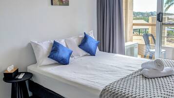 Une chambre d’hôtel avec un lit, des oreillers, une table de chevet avec une boîte de mouchoirs et une télécommande, une fenêtre avec des rideaux et une vue sur des immeubles.