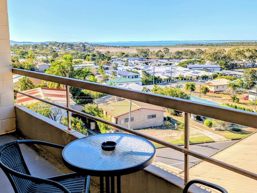 Ein Balkon mit zwei Korbstühlen und einem kleinen runden Tisch mit Blick auf ein Wohngebiet, Häuser und einen klaren Himmel.
