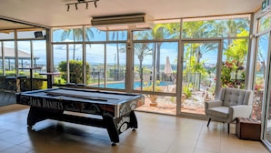A pool table with 'Jack Daniels' branding, large windows with a view of a pool and palm trees, and a chair near the windows. - Lumina Motel (South Gladstone)