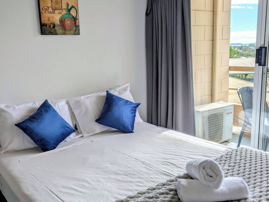Ett hotellrum med en säng, kuddar, ett täcke, en handduk, ett fönster med gardiner och utsikt över omgivningen.