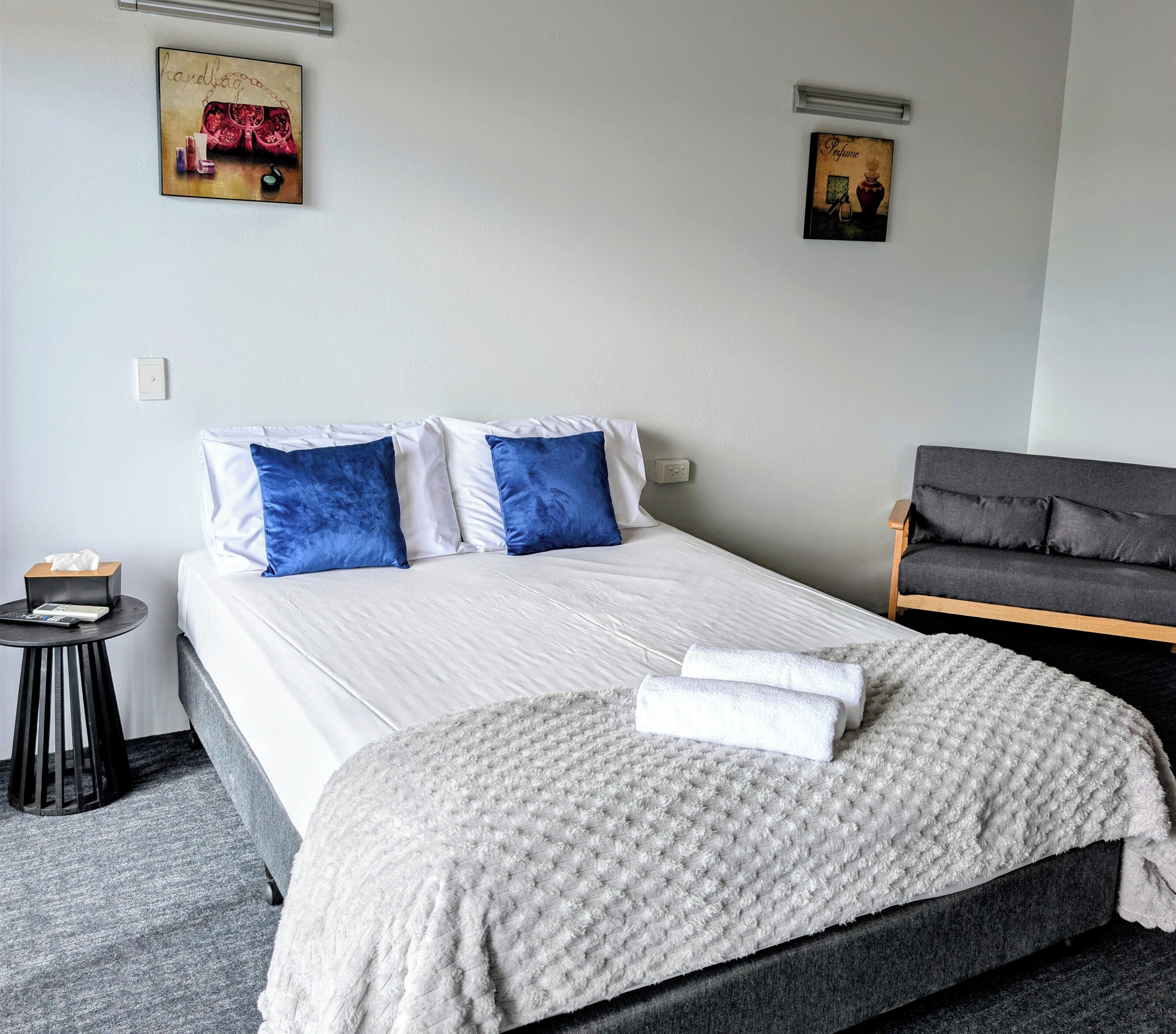 Una cama bien tendida con ropa de cama blanca y azul, un sofá gris y dos cuadros colgados en la pared.