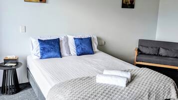 Una cama bien tendida con ropa de cama blanca y azul, un sofá gris y dos cuadros colgados en la pared.
