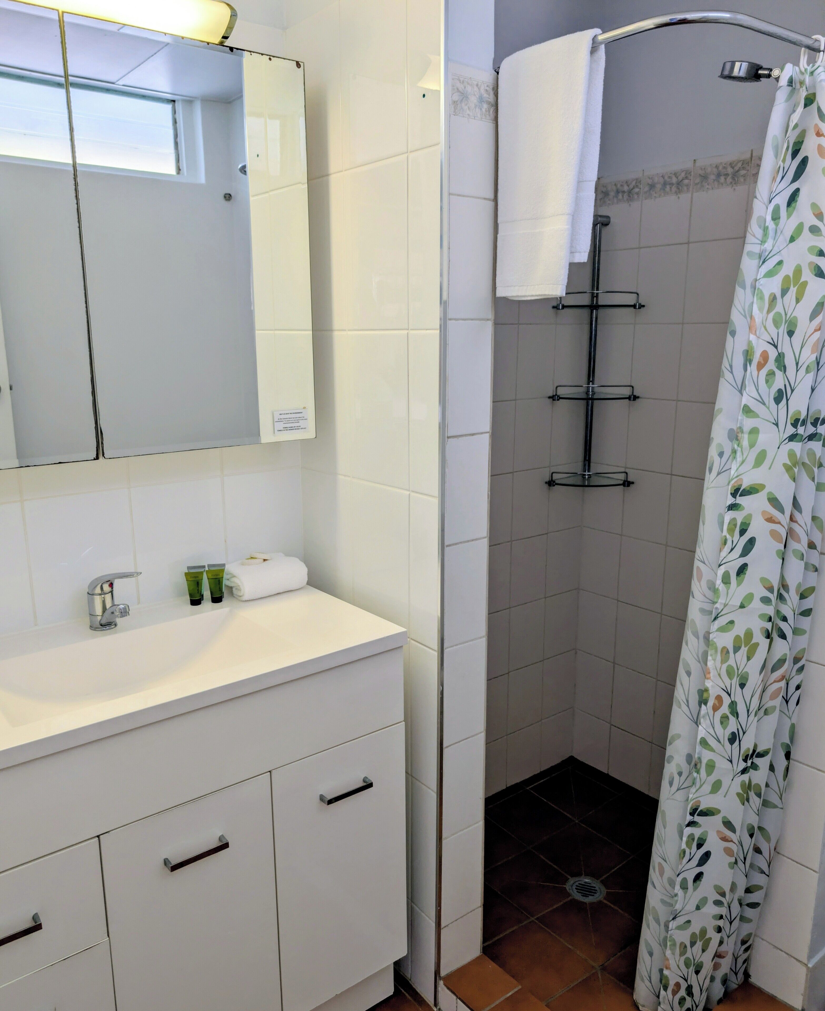 Une salle de bain avec un meuble-lavabo blanc, un miroir, une douche avec un rideau blanc et des murs carrelés.