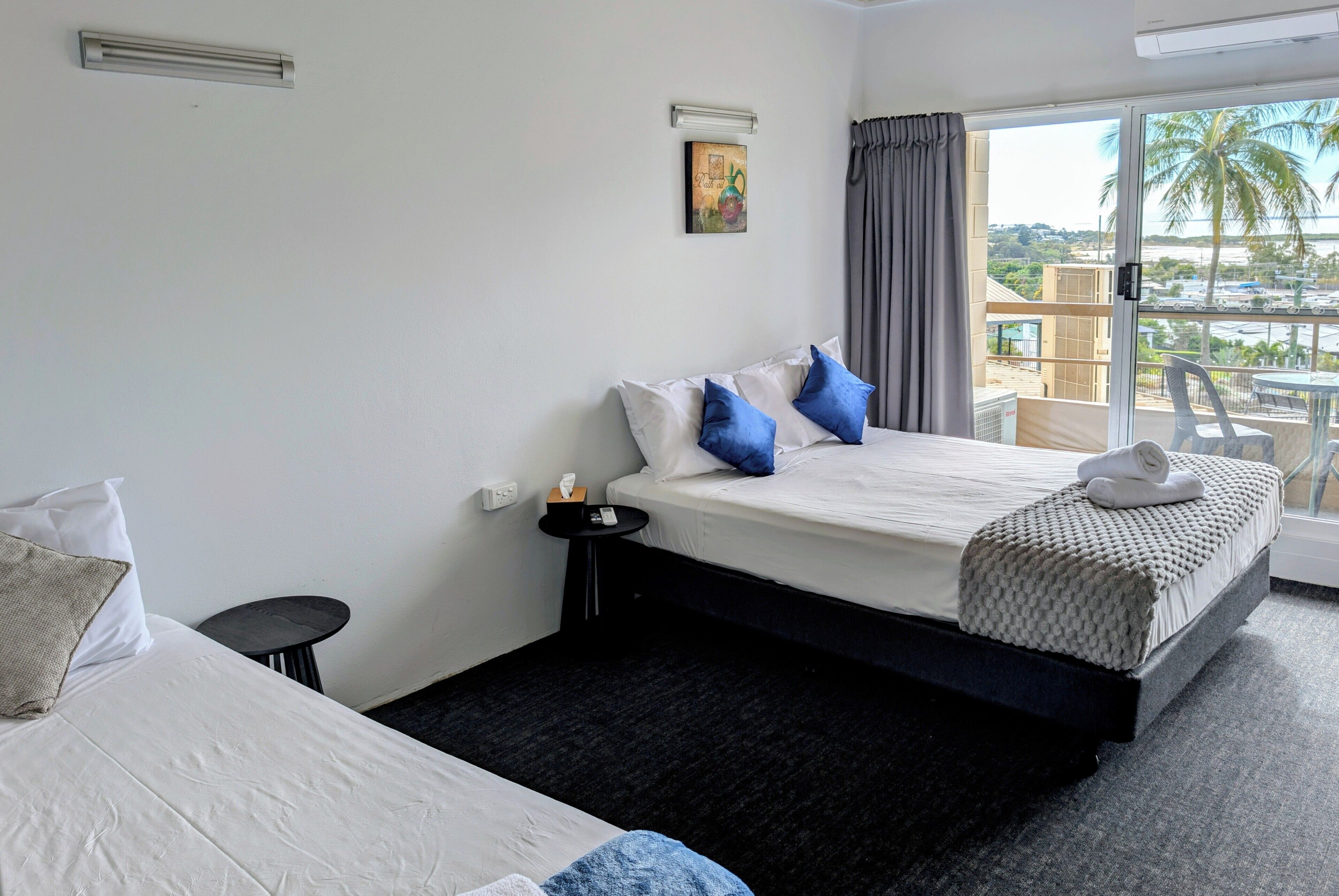 A hotel room with a bed, a small table, a wall-mounted light, a painting, and a balcony with a view.