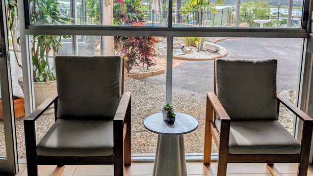 A room with two chairs, a small table, and a view of palm trees and a body of water through large windows. - Lumina Motel (South Gladstone)