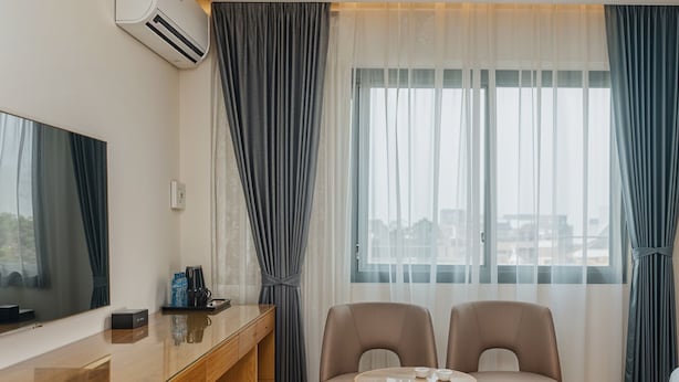 Hotelkamer met een houten bureau, twee lederen fauteuils, een klein rond tafeltje en een groot raam met transparante gordijnen.