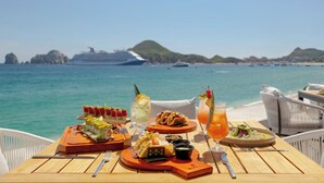 Ein Tisch ist reichhaltig mit Speisen und Getränken gedeckt, mit Blick auf einen Strand und im Hintergrund ein Kreuzfahrtschiff.