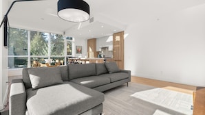 A modern living room with a sectional sofa, a small ottoman, a large window, and a kitchen in the background.