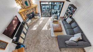 A living room with a stone fireplace, a grey sofa, a bookshelf, and a view through glass doors. - Alpine Pines | Deck | BBQ | Community Amenities (Kings Beach)