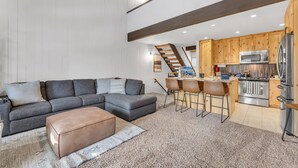 A modern living room with a sectional sofa, ottoman, and a kitchen area with wooden cabinets and stainless steel appliances. - Alpine Pines | Deck | BBQ | Community Amenities (Kings Beach)