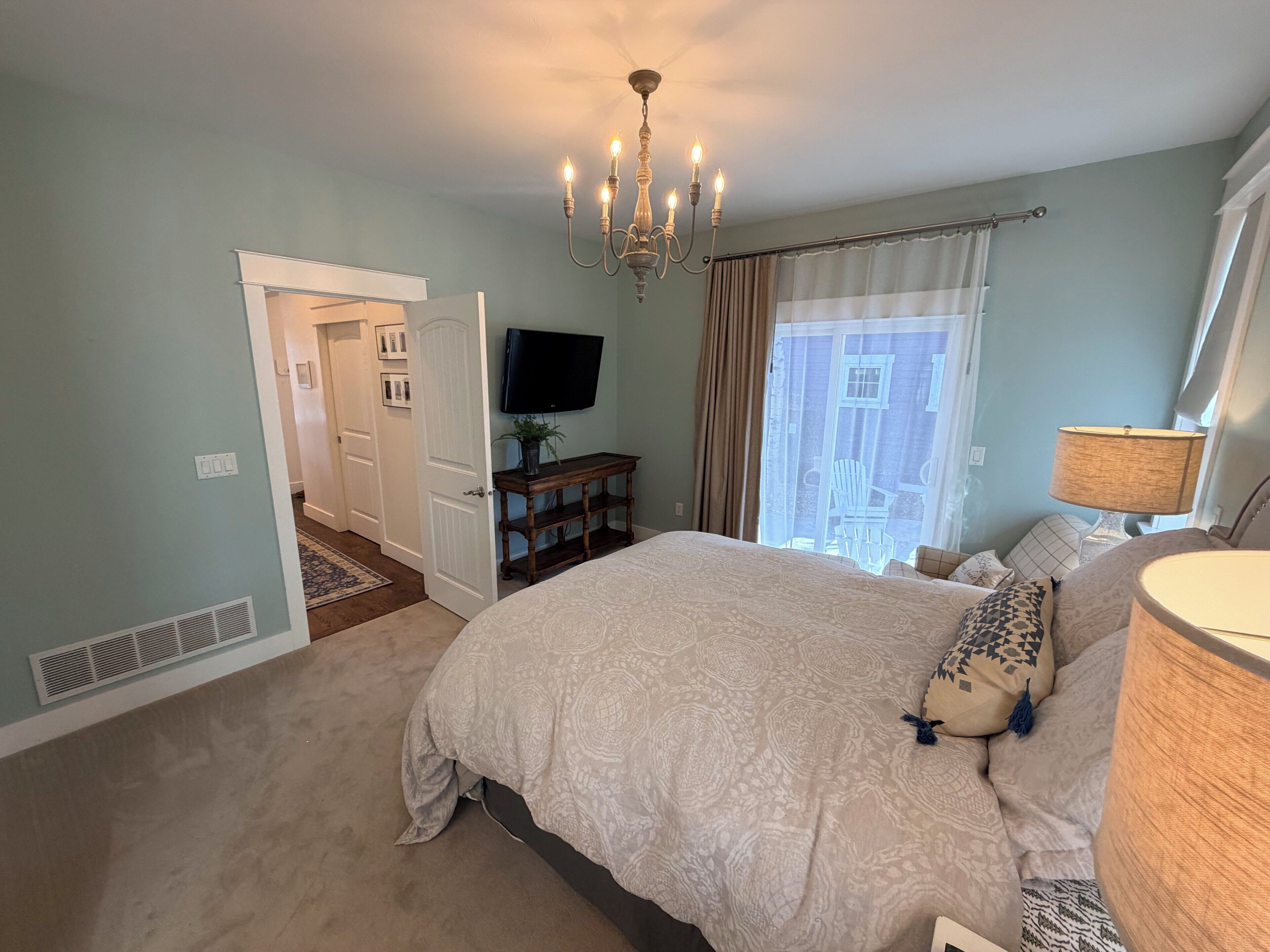 Een slaapkamer met een bed, een televisie, een kroonluchter, een raam met gordijnen en een deur die naar een andere kamer leidt.