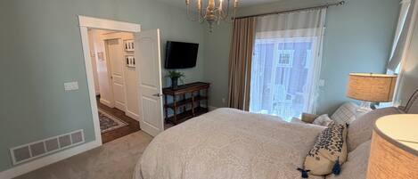 Een slaapkamer met een bed, een televisie, een kroonluchter, een raam met gordijnen en een deur die naar een andere kamer leidt.