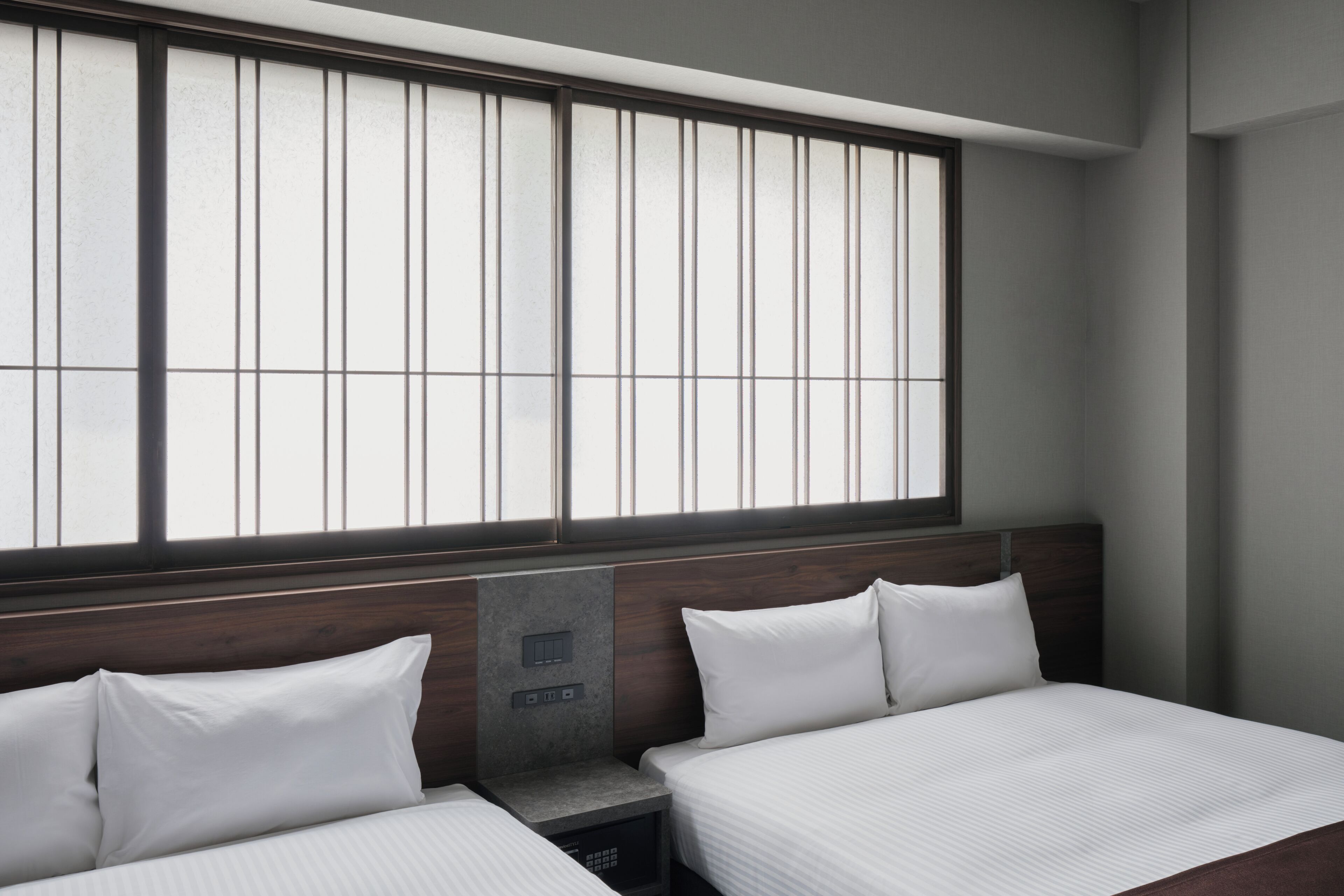 Een hotelkamer met twee bedden, een groot raam met zonwering, een houten hoofdbord en een bedieningspaneel aan de muur.