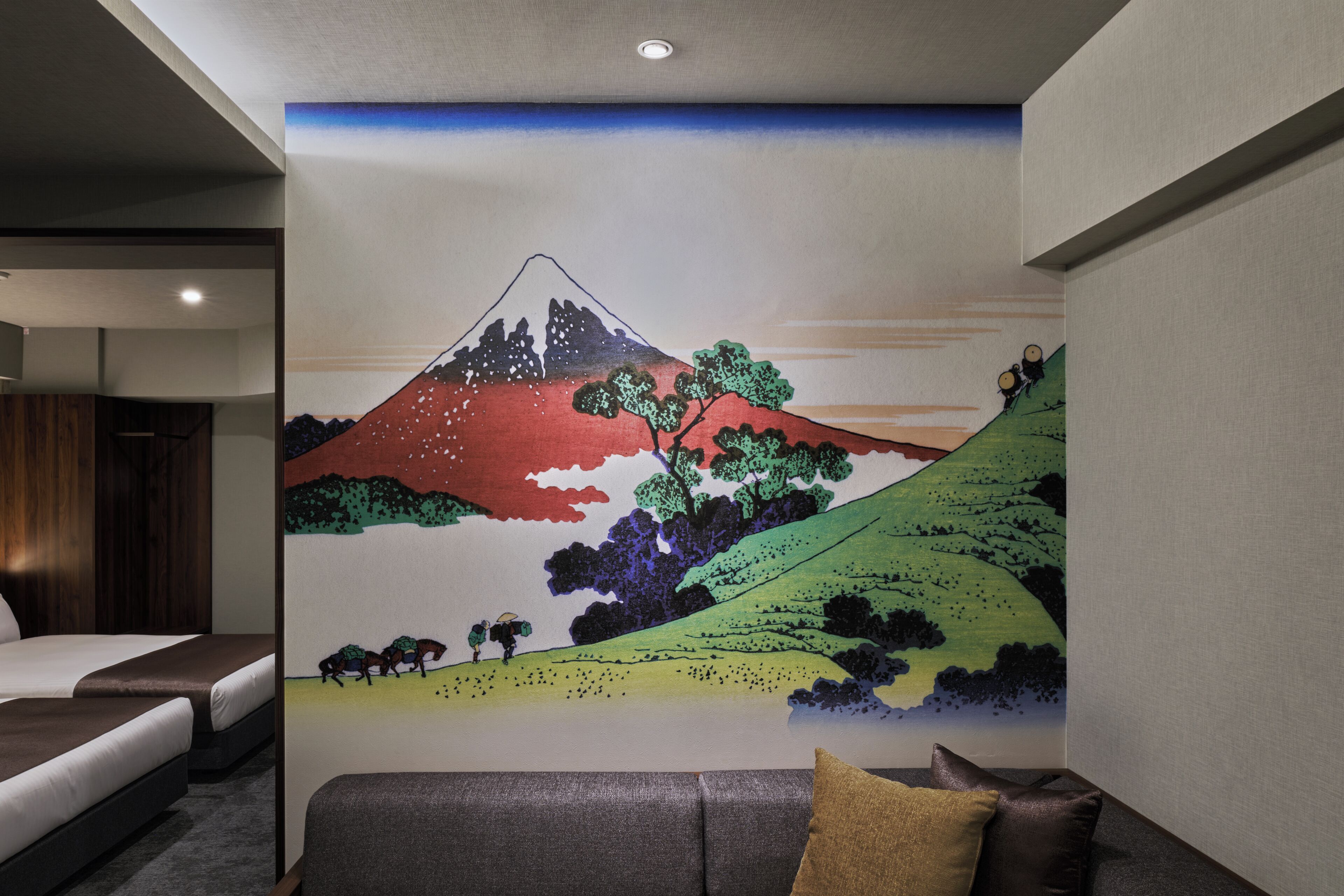A hotel room with a large wall mural of a mountain landscape, a bed, a sofa, and a small table.