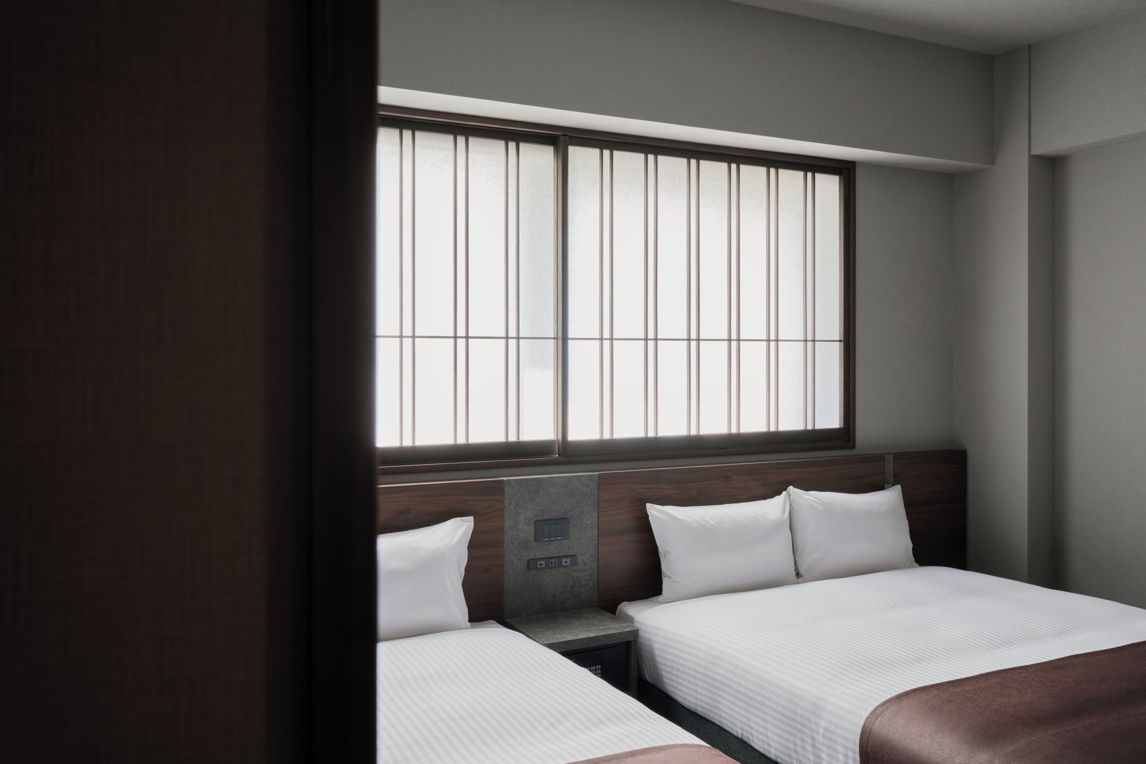 A hotel room with two beds, a window with blinds, a bedside table, and a control panel.